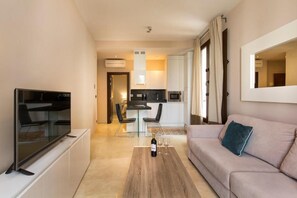 A modern living room with a flat-screen TV, a sofa, a coffee table with a bottle and glasses, and a kitchen in the background. - New Listing (Granada)