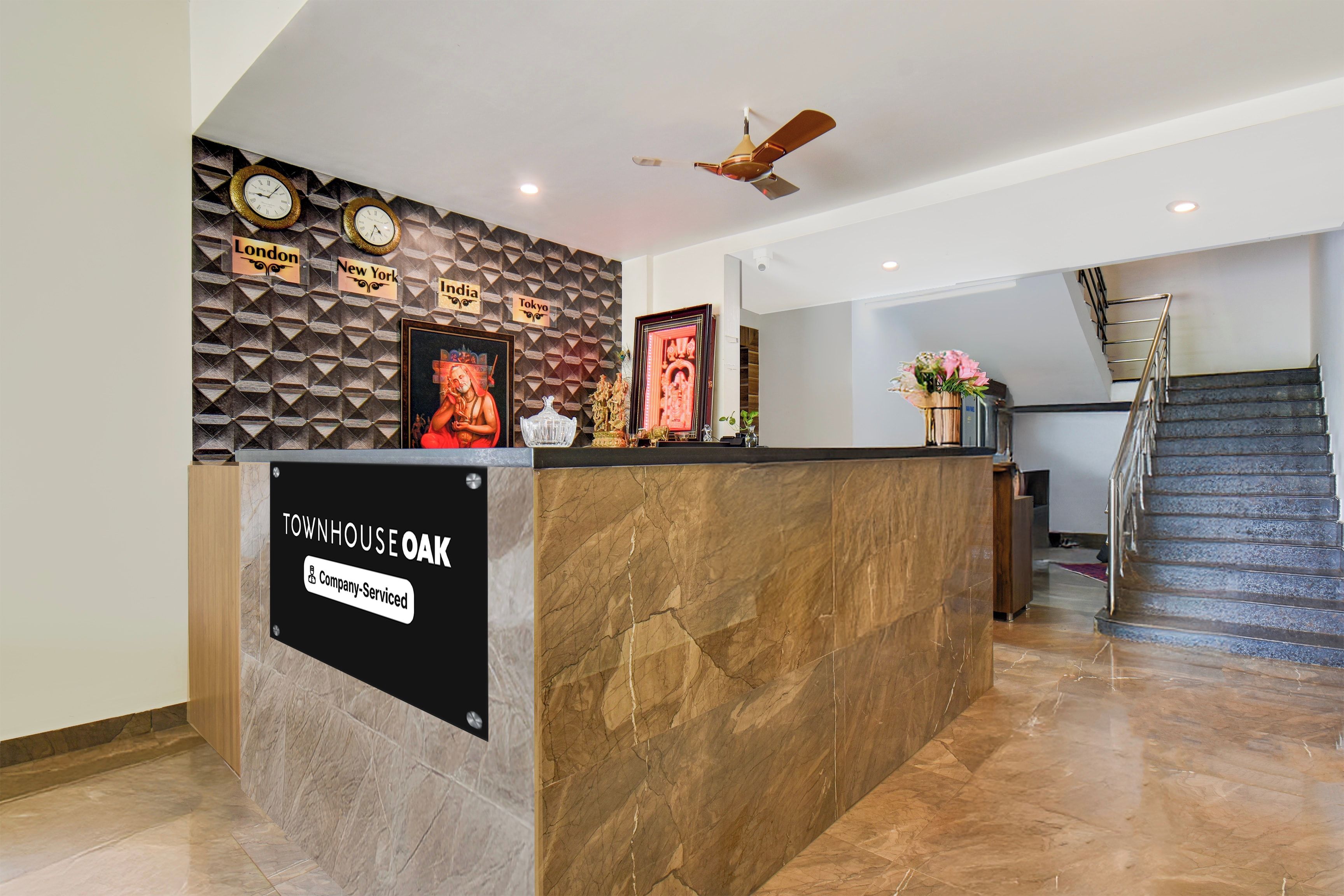 Un mostrador de recepción con un cartel que indica servicio de la empresa, una pared con relojes y nombres de ciudades, y una escalera al fondo.