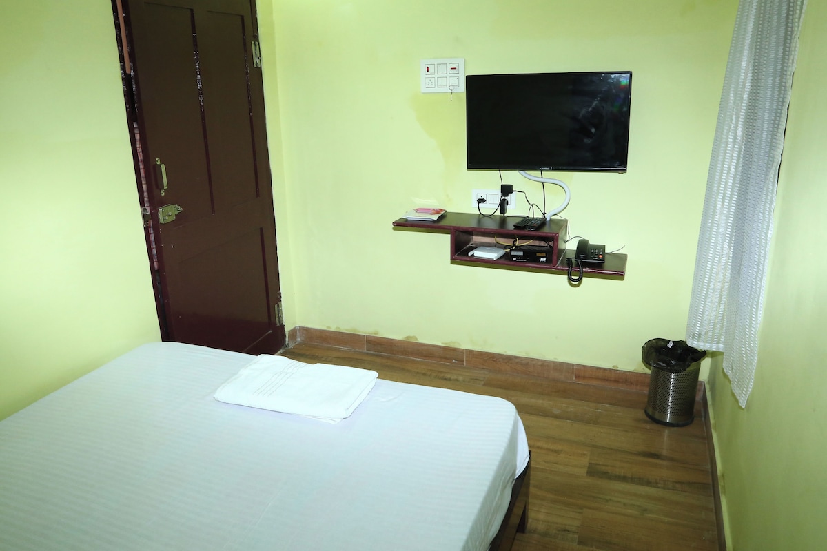 A hotel room with a bed, a TV mounted on the wall, a small shelf with items, and a trash bin.