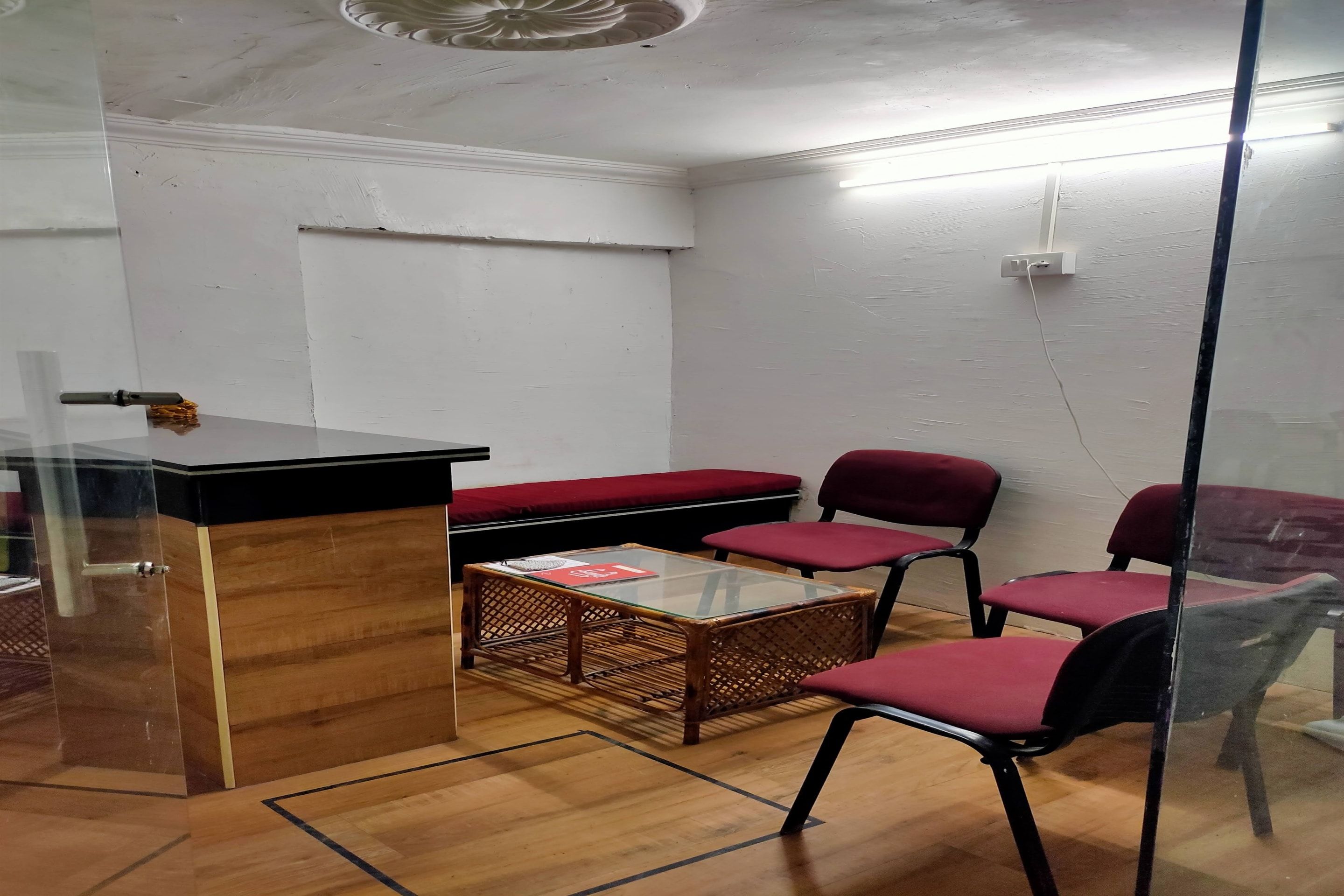 Huoneessa on lasiseinä, puinen työpöytä, punaisella kankaalla verhoiltu penkki ja kaksi punaista tuolia.