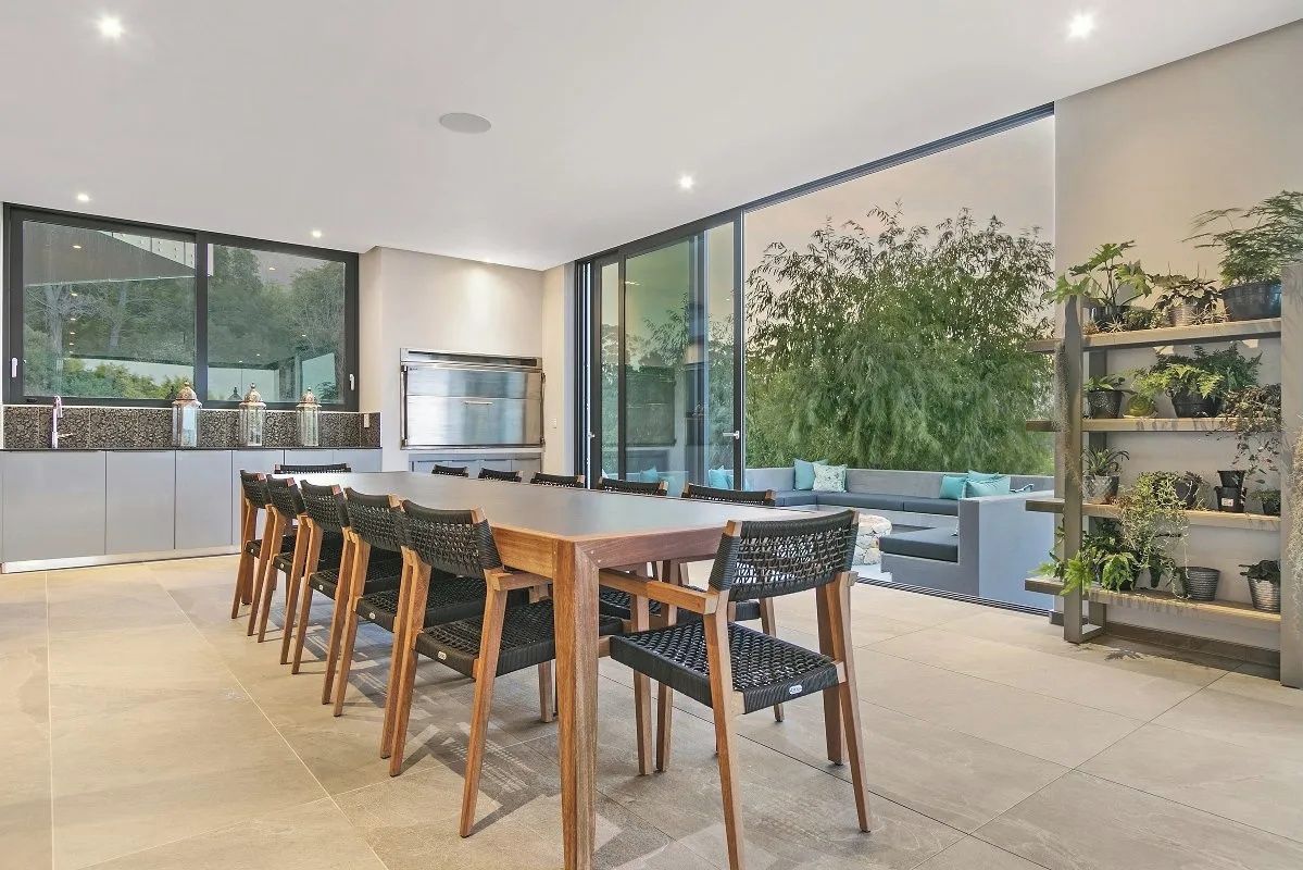 A modern kitchen with a dining area, large windows, and a view of greenery.