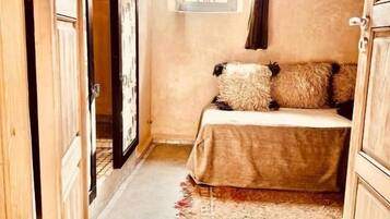 Un couloir étroit mène à une chambre à coucher qui contient un lit, une fenêtre ornée de ferronnerie décorative et une porte en bois.