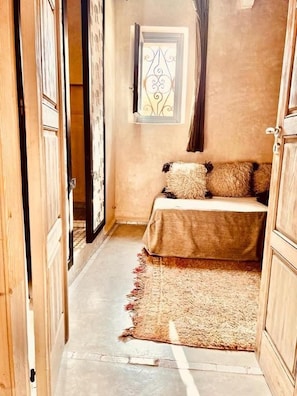A narrow hallway leading to a bedroom with a bed, a window with decorative ironwork, and a wooden door. - Maison D'Hôtes DAR LES SIRENES (Essaouira)