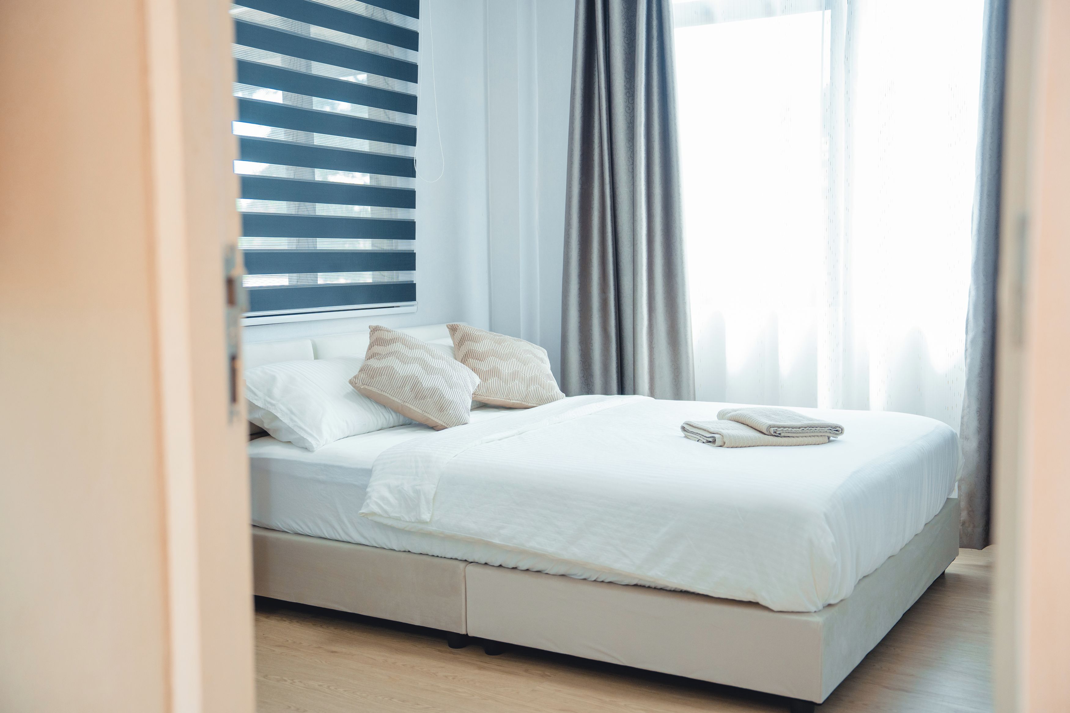 Una cama bien hecha con ropa blanca y dos almohadas, una ventana con persianas y una cortina.