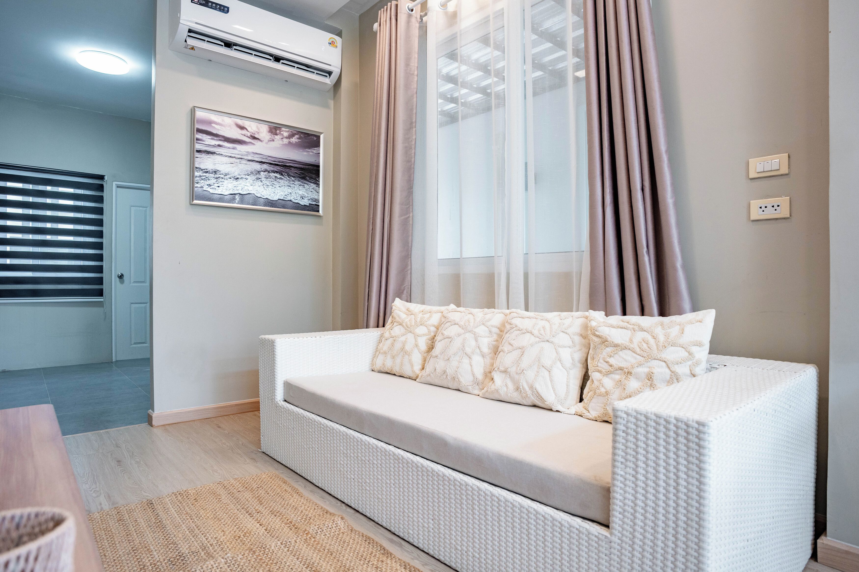 A white sofa with patterned cushions, a window with sheer curtains, an air conditioning unit, and a framed picture on the wall.