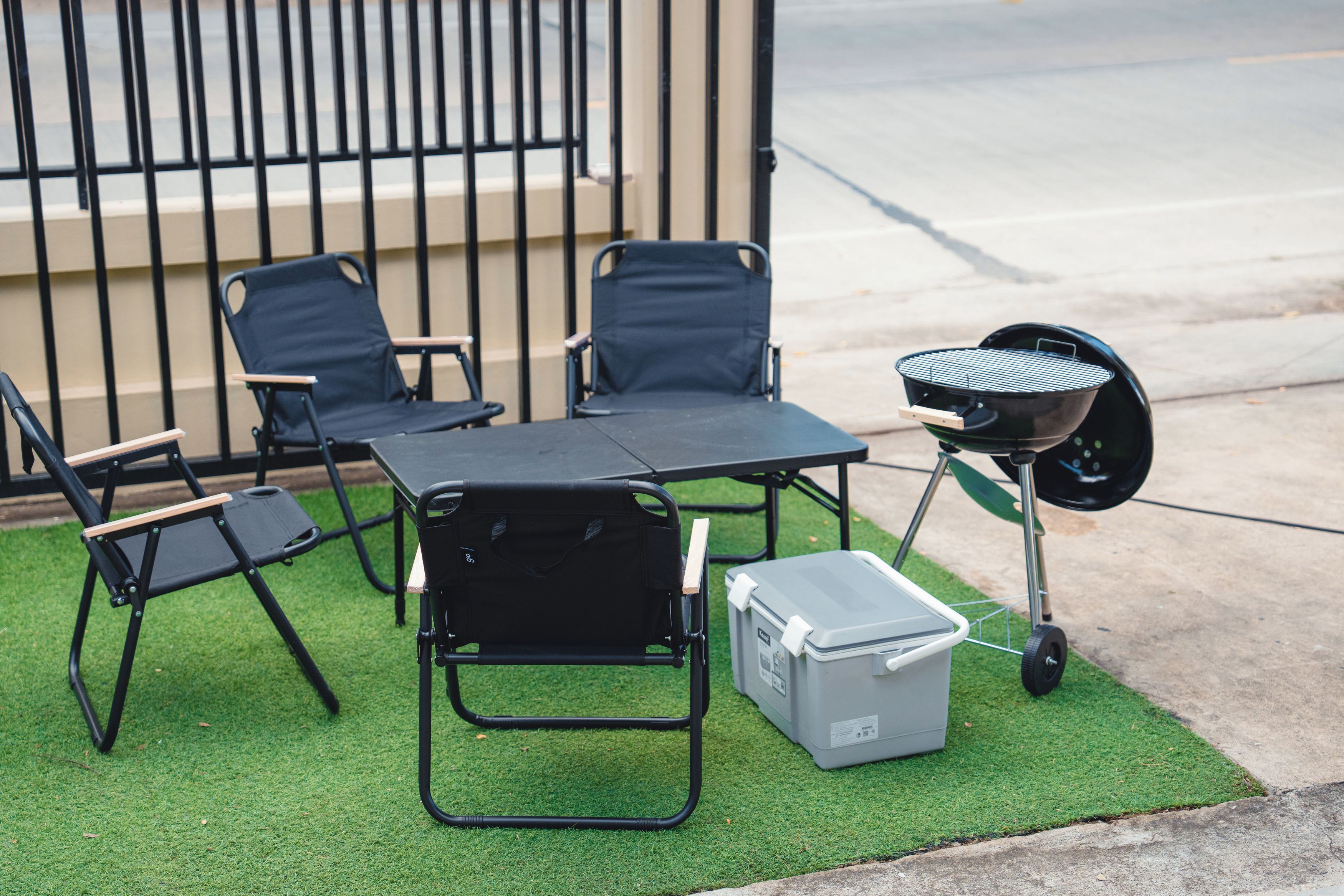 A portable grill set up on artificial grass with a table and chairs, a cooler, and a wheelbarrow.