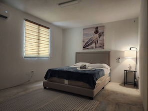 A bedroom with a bed, a window with blinds, a painting of a surfer, a bedside lamp, and a rug. - Nice Place to Relax Leve I (Fajardo)
