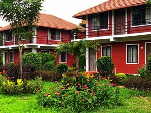 A red building with multiple windows and balconies, surrounded by a well-maintained garden with various plants and flowers. - Streamedge Resort (Sakleshpur)