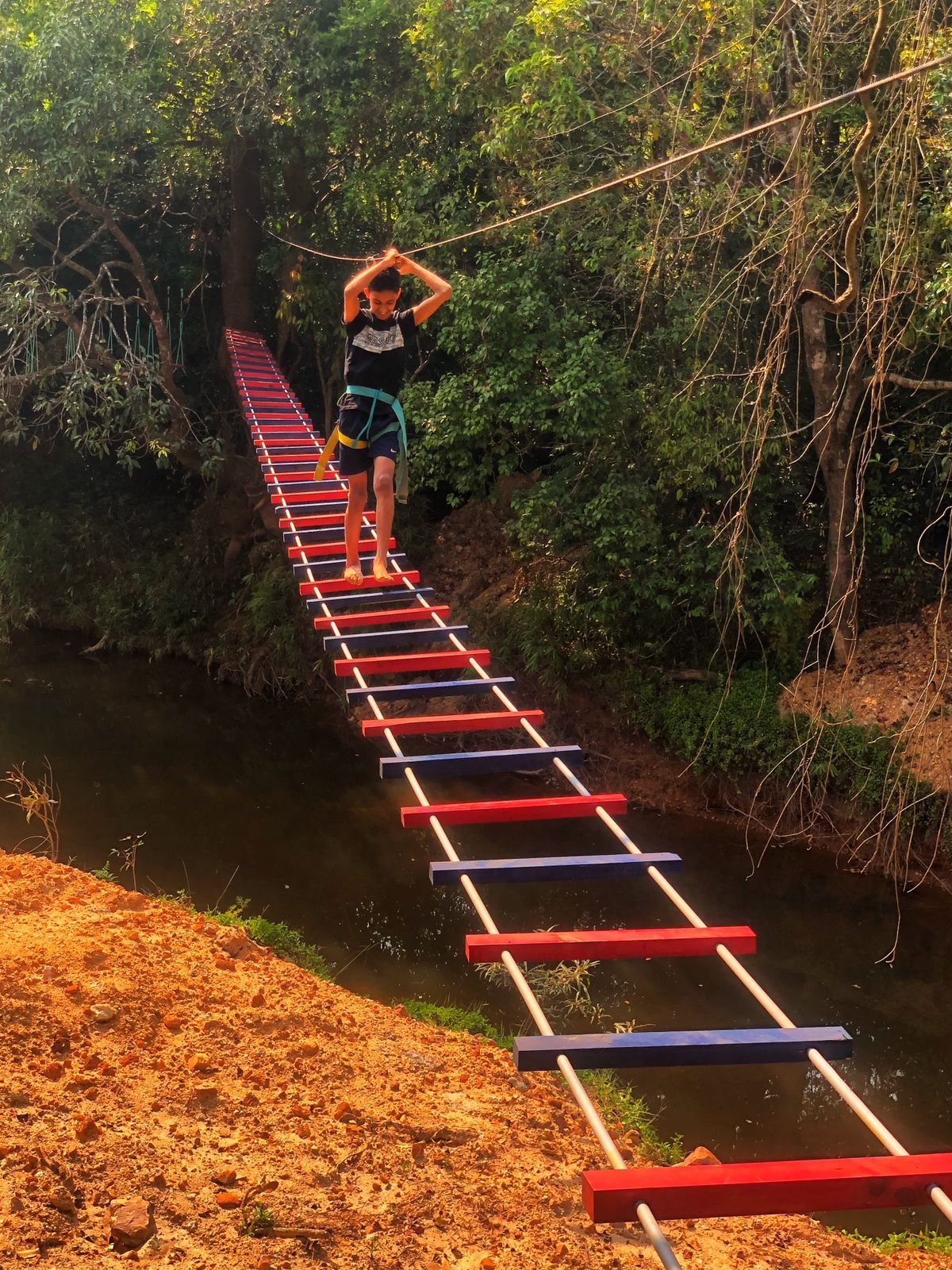 A person walking on a suspended bridge over a river, surrounded by greenery.