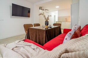 Una sala moderna con un sofá rojo, una mesa de comedor con sillas, un televisor de pantalla plana en la pared y una cocina al fondo.