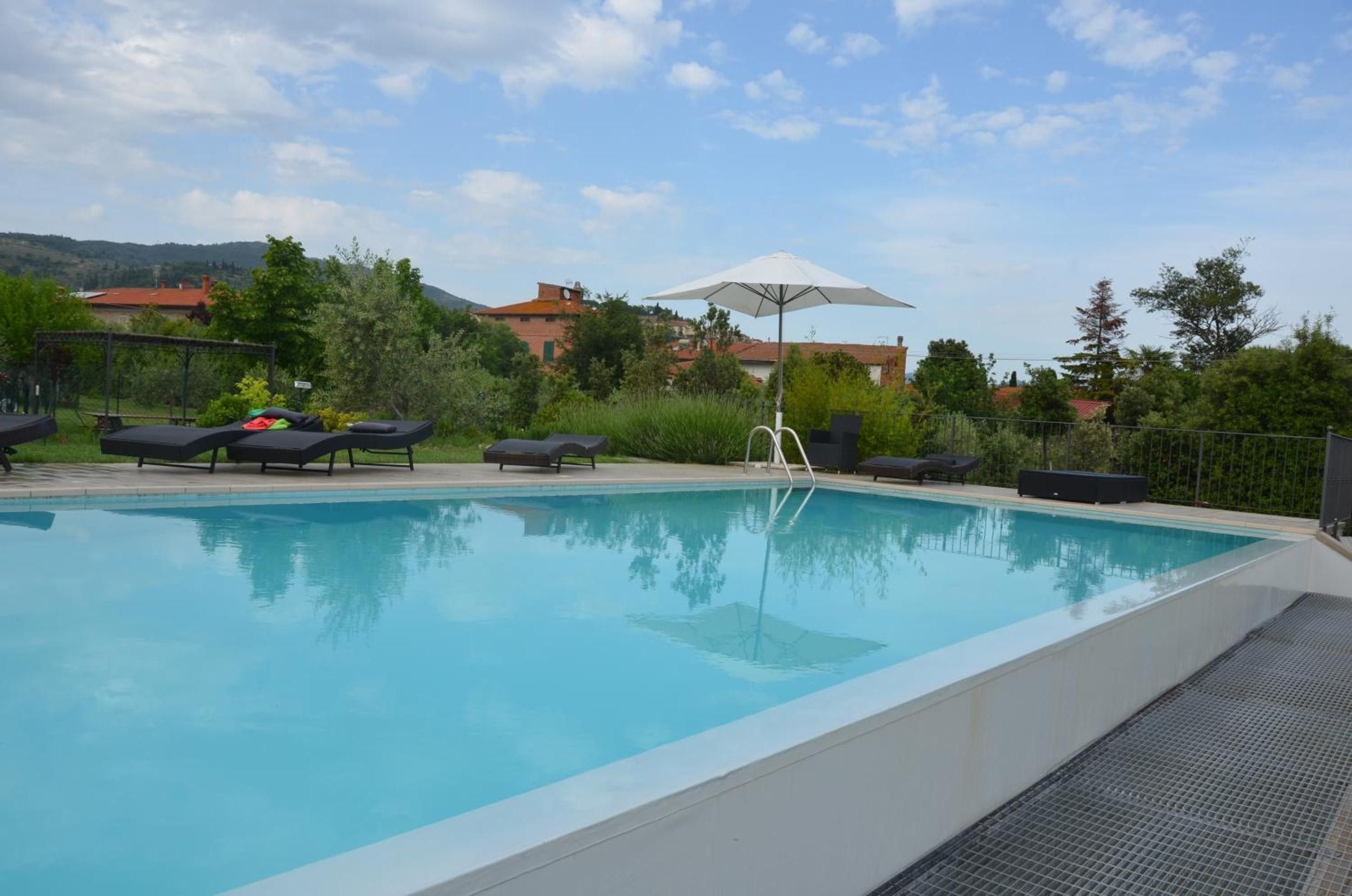 A clear swimming pool with lounge chairs and an umbrella, surrounded by greenery and buildings in the background.