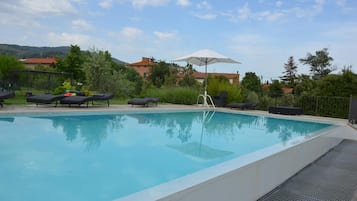 A clear swimming pool with lounge chairs and an umbrella, surrounded by greenery and buildings in the background.