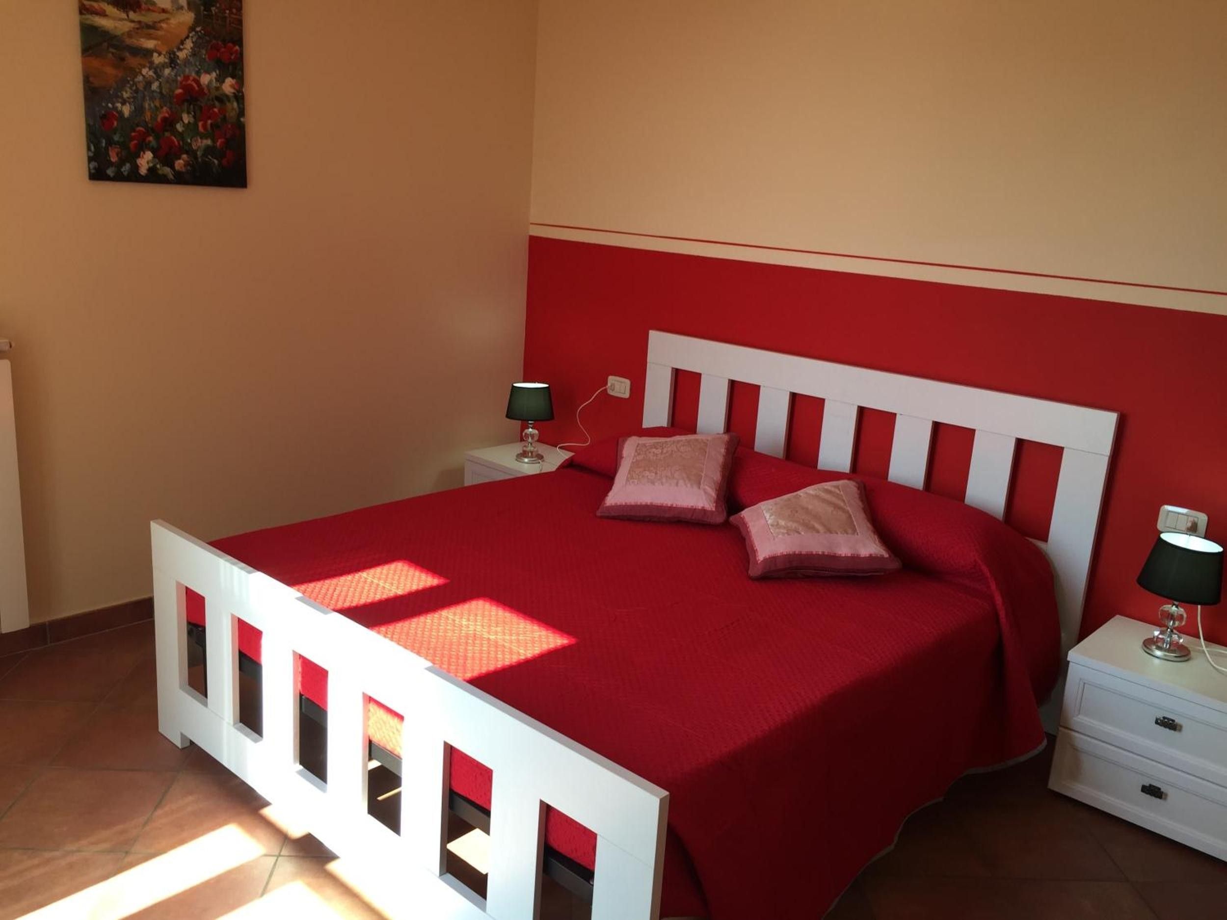 A bedroom with a red bedspread, white headboard, two bedside tables with lamps, and a painting on the wall.