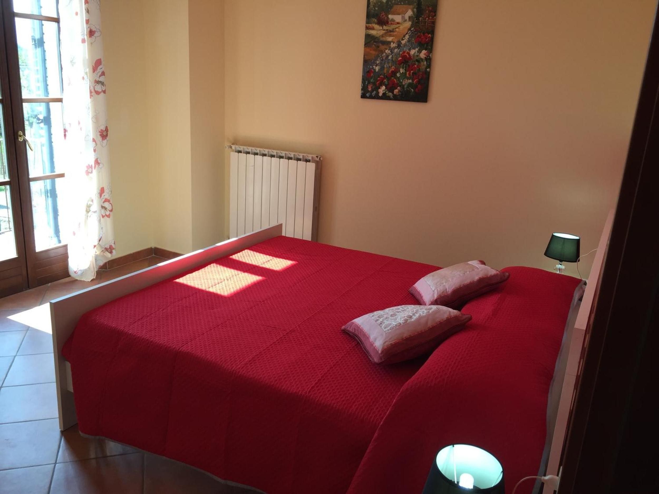 A bedroom with a red bedspread, two pillows, a radiator, a painting on the wall, and a window with curtains.