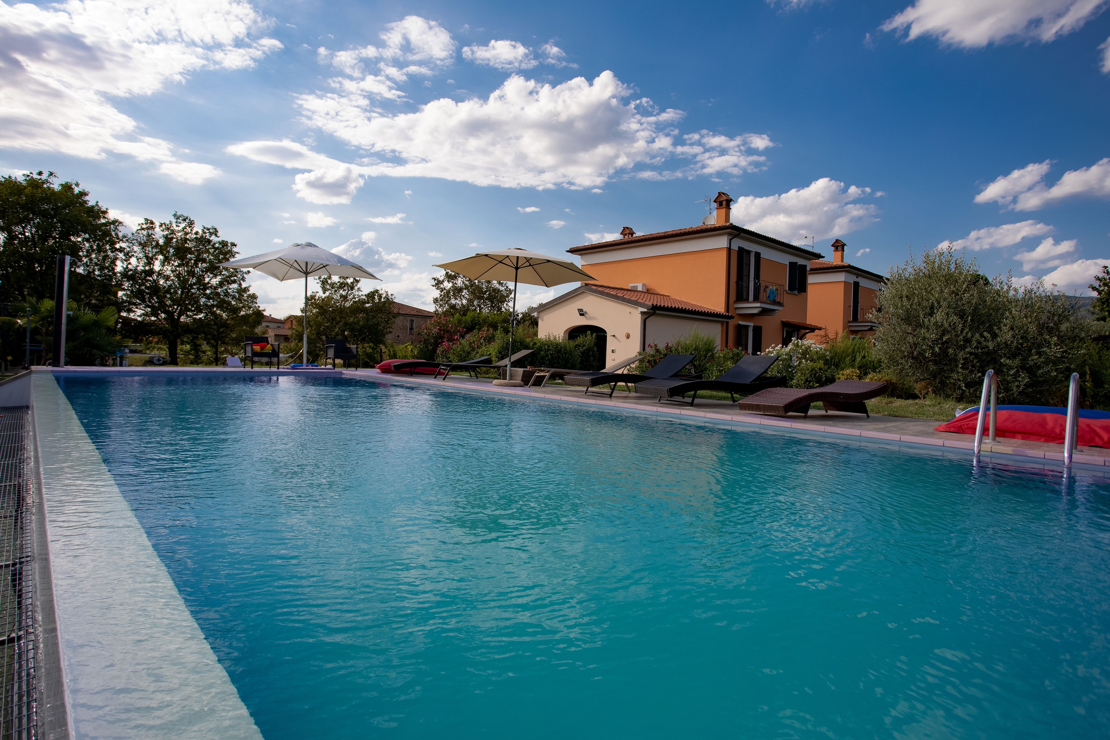 A swimming pool with lounge chairs and umbrellas, surrounded by a building with a tiled roof.