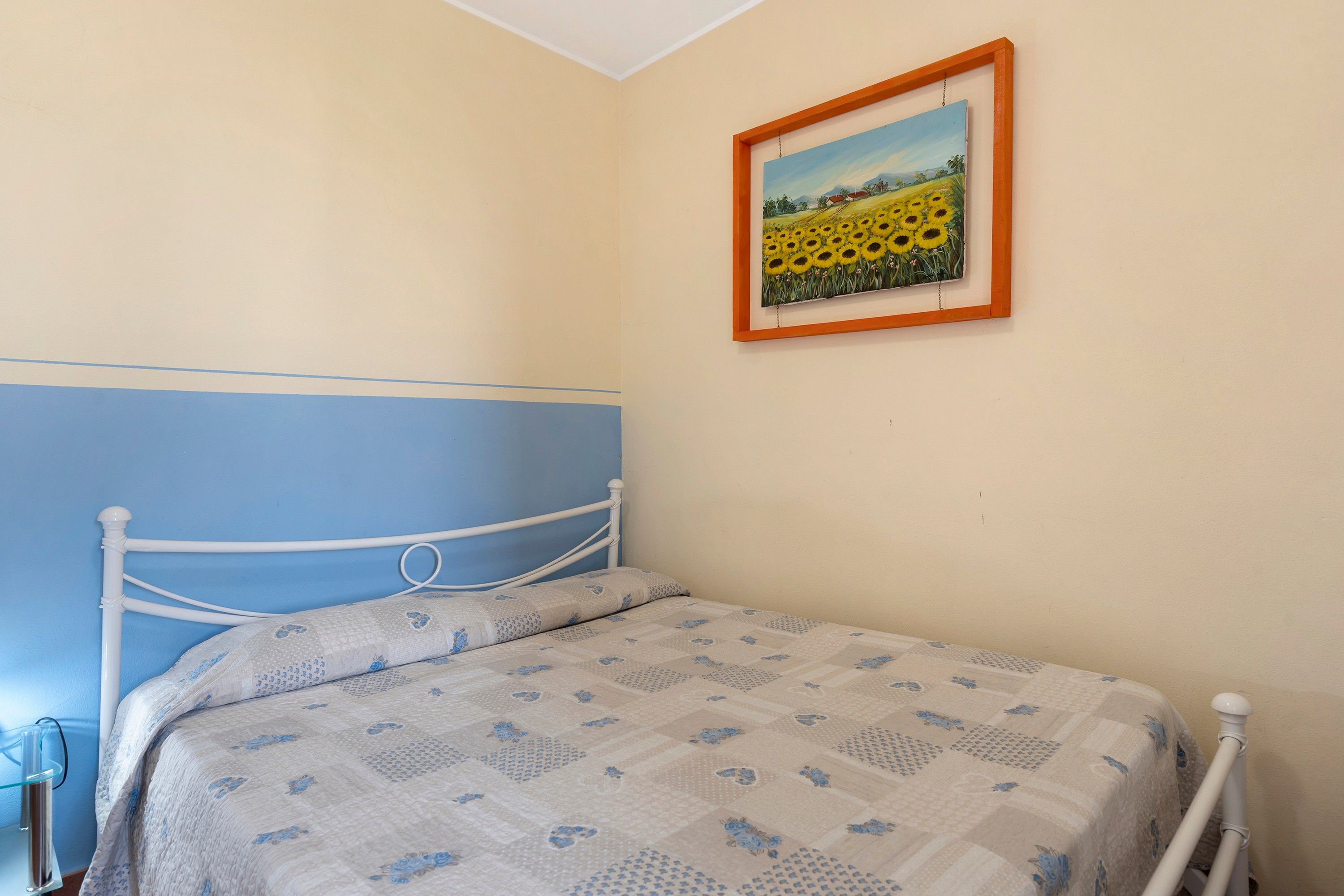 A bedroom with a single bed, a blue headboard, and a framed painting of sunflowers on the wall.
