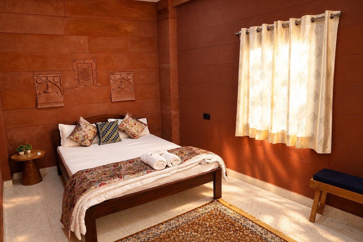 A bedroom with a bed, pillows, a bedside table with plants, a bench, and a patterned rug.
