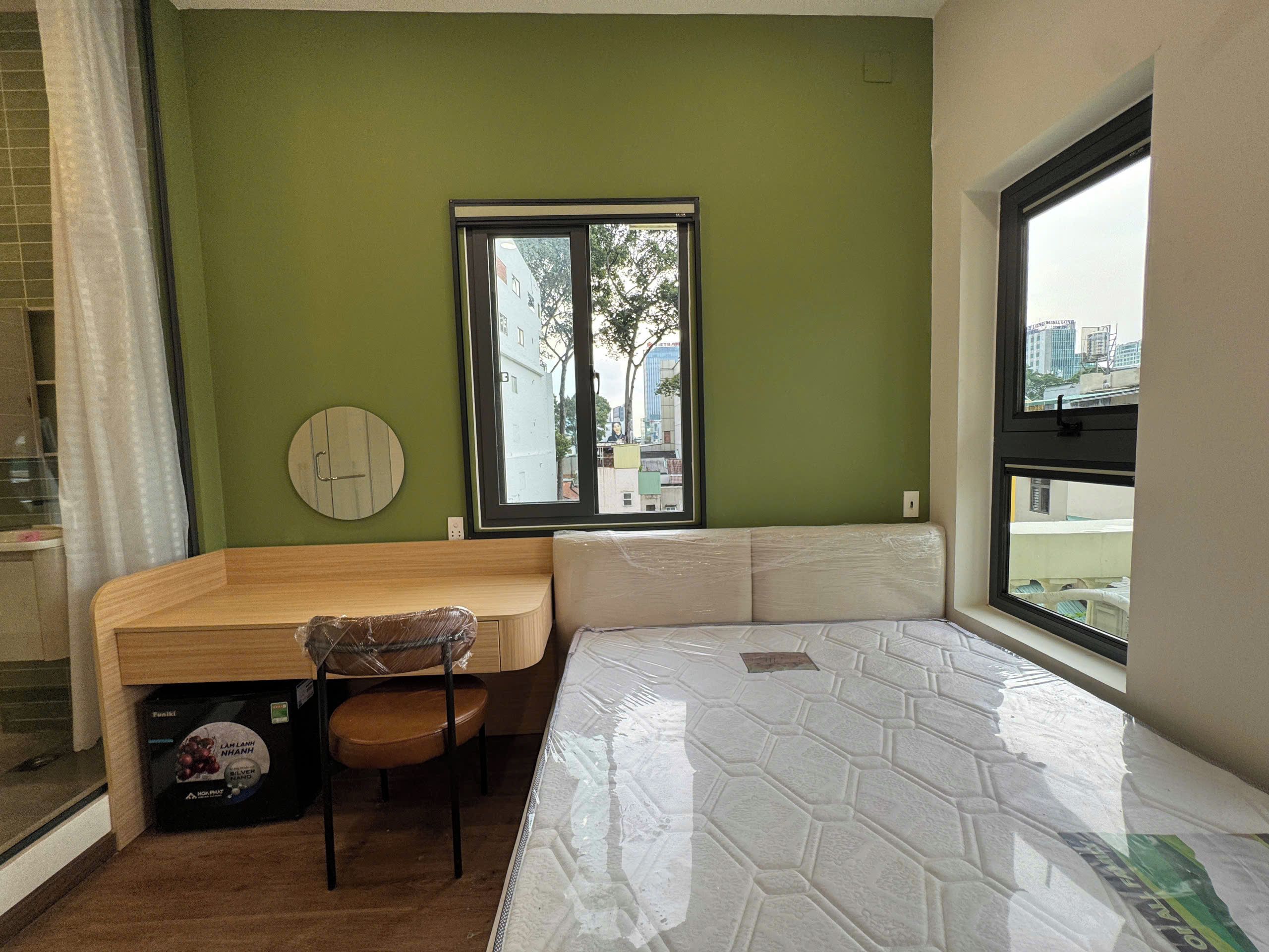 A room with a bed, a wooden desk, a chair, and a window with a view of buildings.