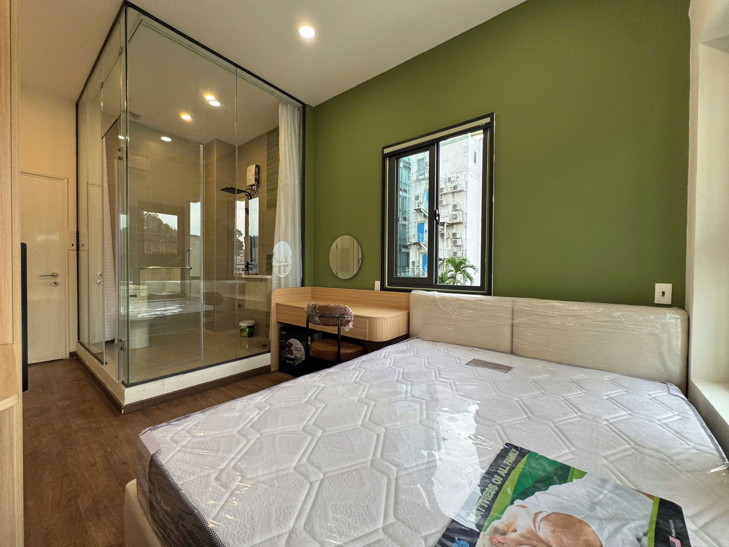 A bedroom with a glass shower enclosure, a window with a view of buildings, and a mattress with a protective cover.