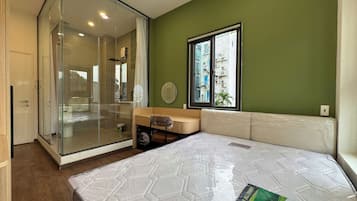 Un dormitorio con cabina de ducha de vidrio, una ventana con vistas a edificios y un colchón con cubierta protectora.