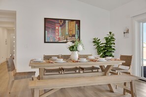 A dining area with a wooden table set for four, a large painting on the wall, and a potted plant. - The Reserve | Modern 5BR Henderson Pool Home (Henderson)
