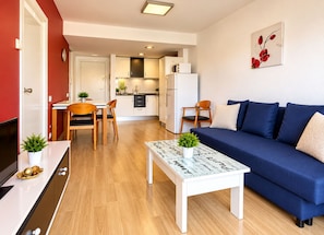 A modern living room with a blue sofa, a white coffee table with a plant, and a kitchen in the background. - Yamila (Salou)