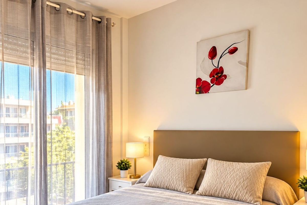 A hotel room with a large bed, a nightstand with a lamp, a wall-mounted artwork, and a view of the outside.