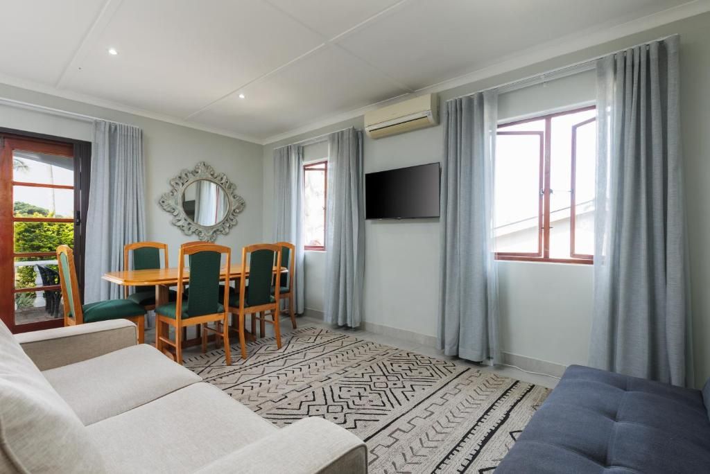A modern living room with a sofa, a dining table with chairs, a TV mounted on the wall, and large windows with curtains.