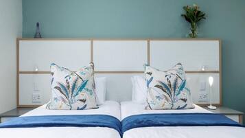 Quarto de hotel com duas camas, cabeceira de madeira, mesa de cabeceira com abajur e um vaso de flores na parede.