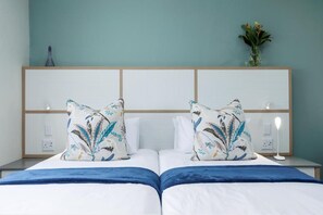 A hotel room with two beds, a wooden headboard, a nightstand with a lamp, and a vase with flowers on the wall.