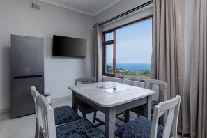 A room with a dining table and chairs, a flat-screen TV, a refrigerator, and a window with a view of the sea.