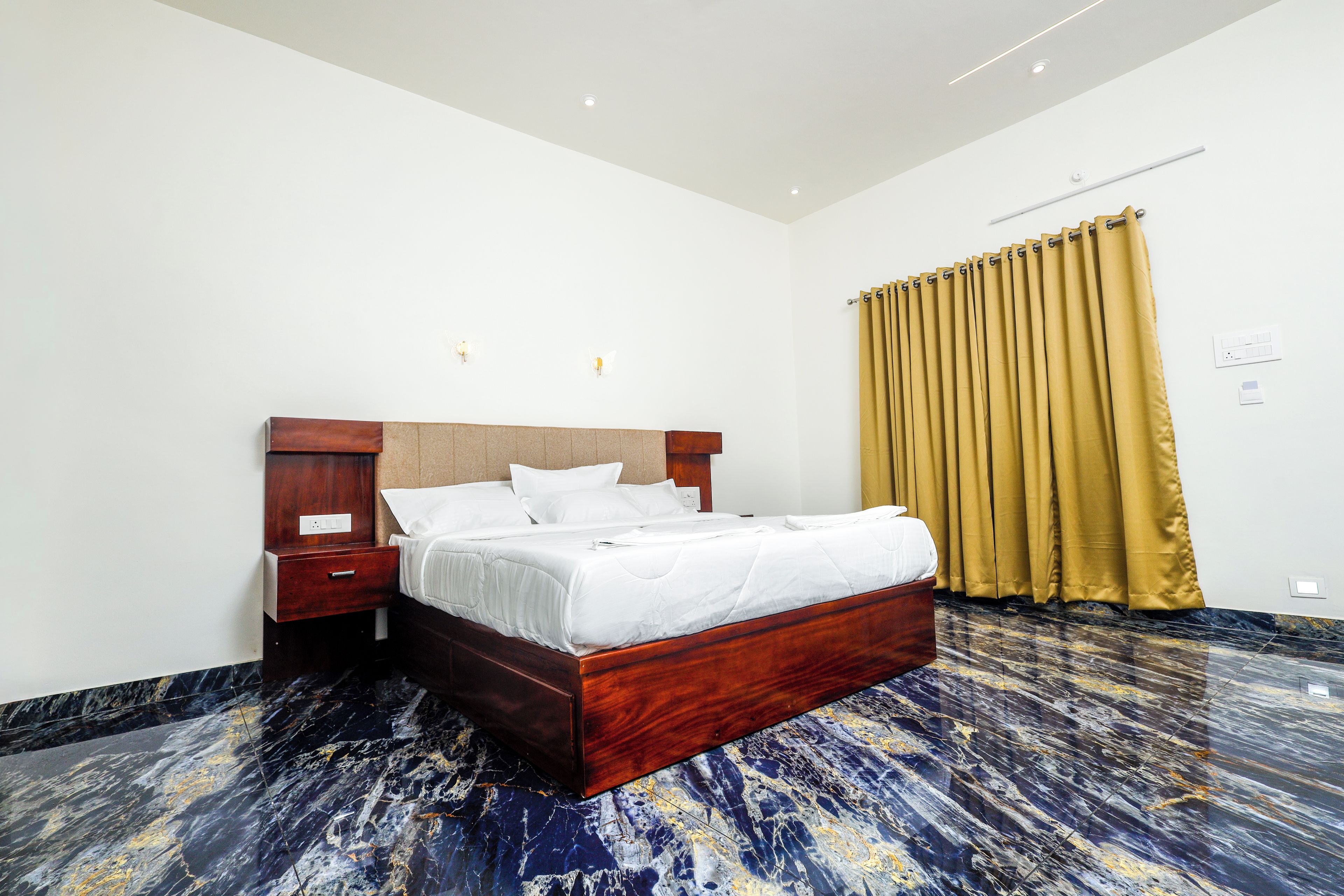 Quarto de hotel com cama de madeira, mesa de cabeceira, luminária na parede e cortina.