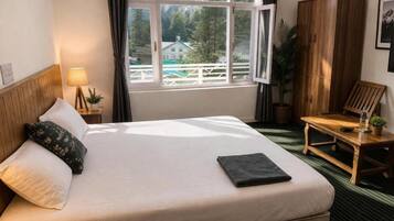 Une chambre d’hôtel avec un lit, une fenêtre avec des rideaux, une armoire en bois, une petite table avec une lampe, et une vue sur une montagne et une maison.