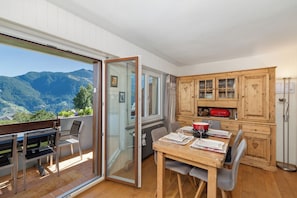 Un comedor con una mesa dispuesta para dos, un armario de madera con puertas de cristal, y vistas a montañas a través de un amplio ventanal corredizo.