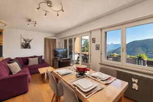 Un salón moderno con sofá, televisor, mesa de comedor y ventanas amplias con vistas a la montaña.