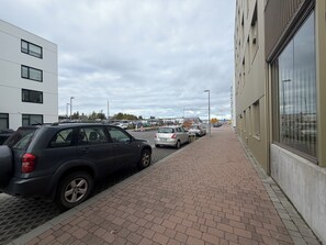 En parkeringsplats med bilar som står uppradade längs en asfalterad gångväg intill en byggnad med stora fönster.