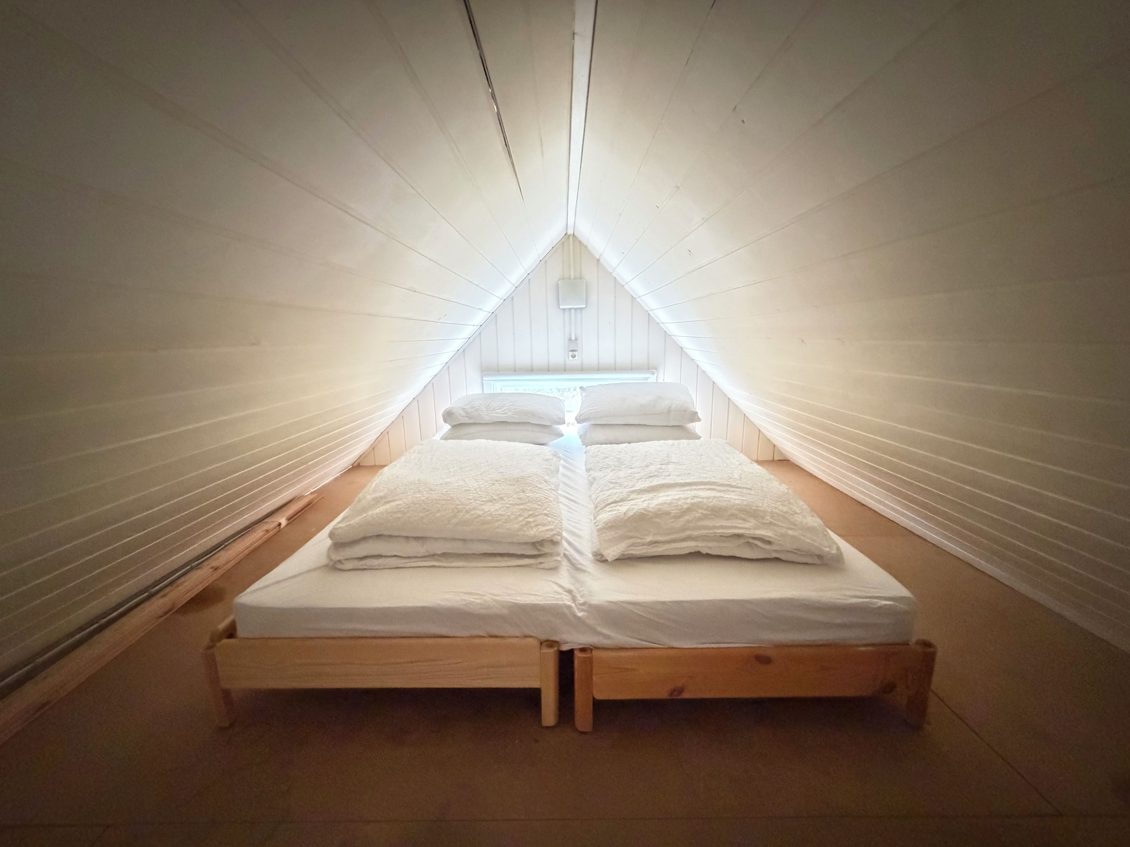 Quarto sótão pequeno e triangular com cama de madeira e roupa de cama branca.