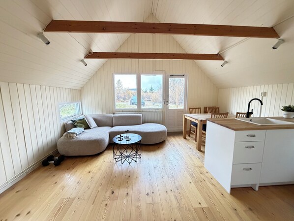 A loft-style room with a wooden ceiling, a dining area, a kitchen with a sink, and a large window providing a view of the outdoors.