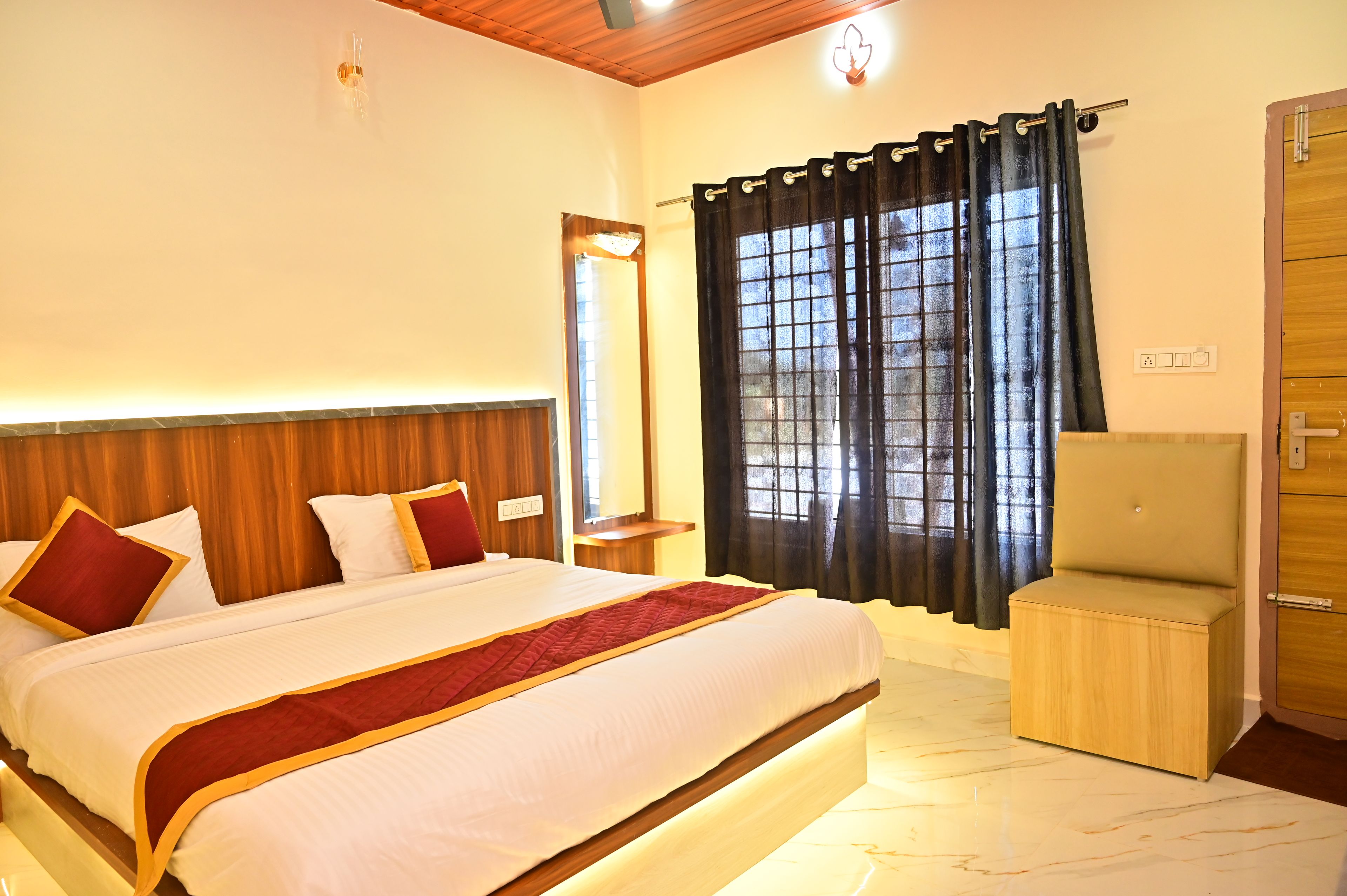 Una habitación de hotel con una cama, un cabecero de madera, una mesita de noche, una silla, un ventanal con cortinas y una puerta.