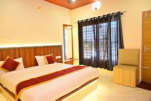 Quarto de hotel com cama, cabeceira de madeira, mesa de cabeceira, cadeira, janela com cortinas e porta.