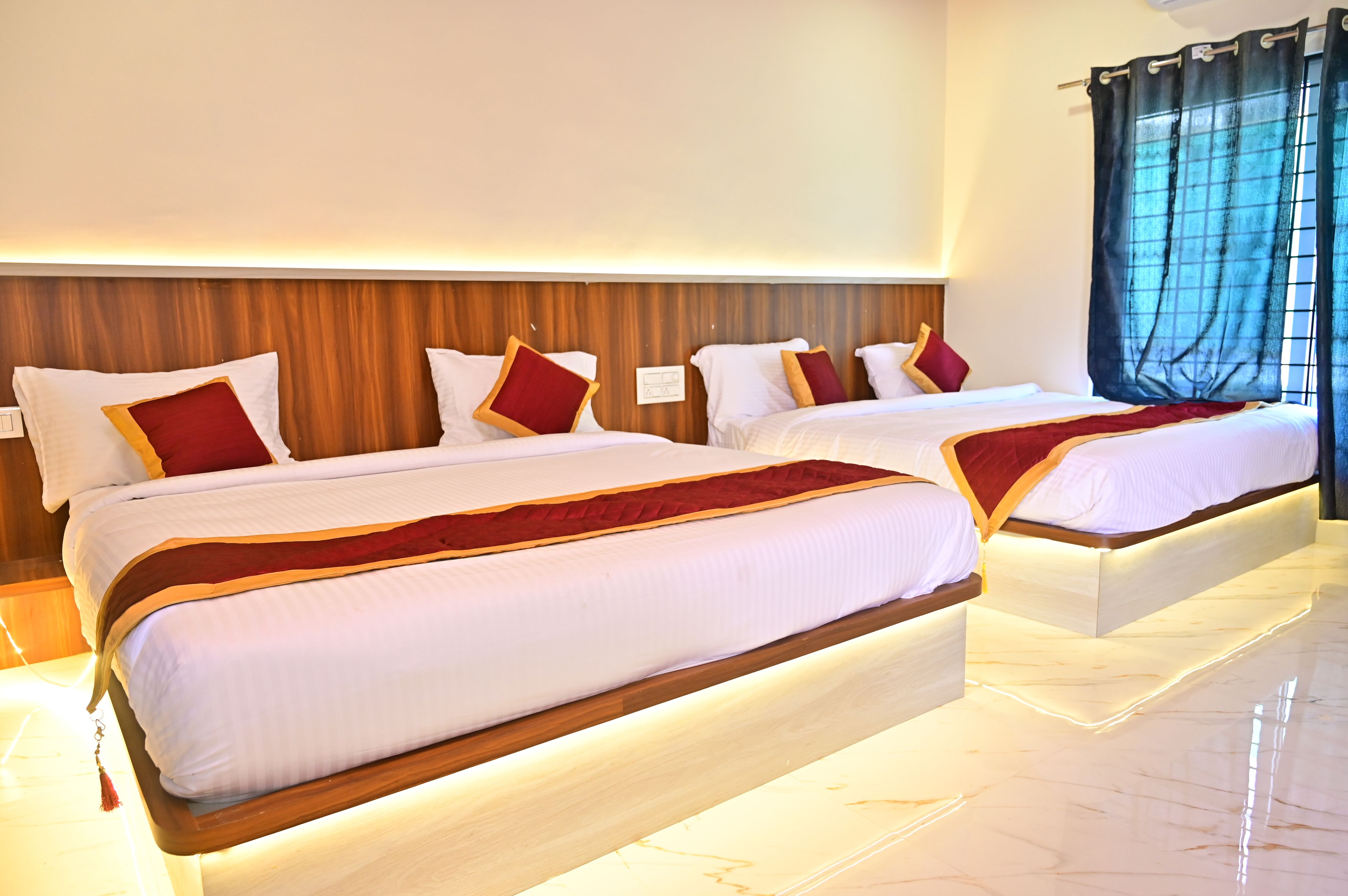 Une chambre d’hôtel avec deux lits, des têtes de lit en bois, du linge de lit blanc, des coussins décoratifs rouges et une grande fenêtre avec des rideaux bleus.