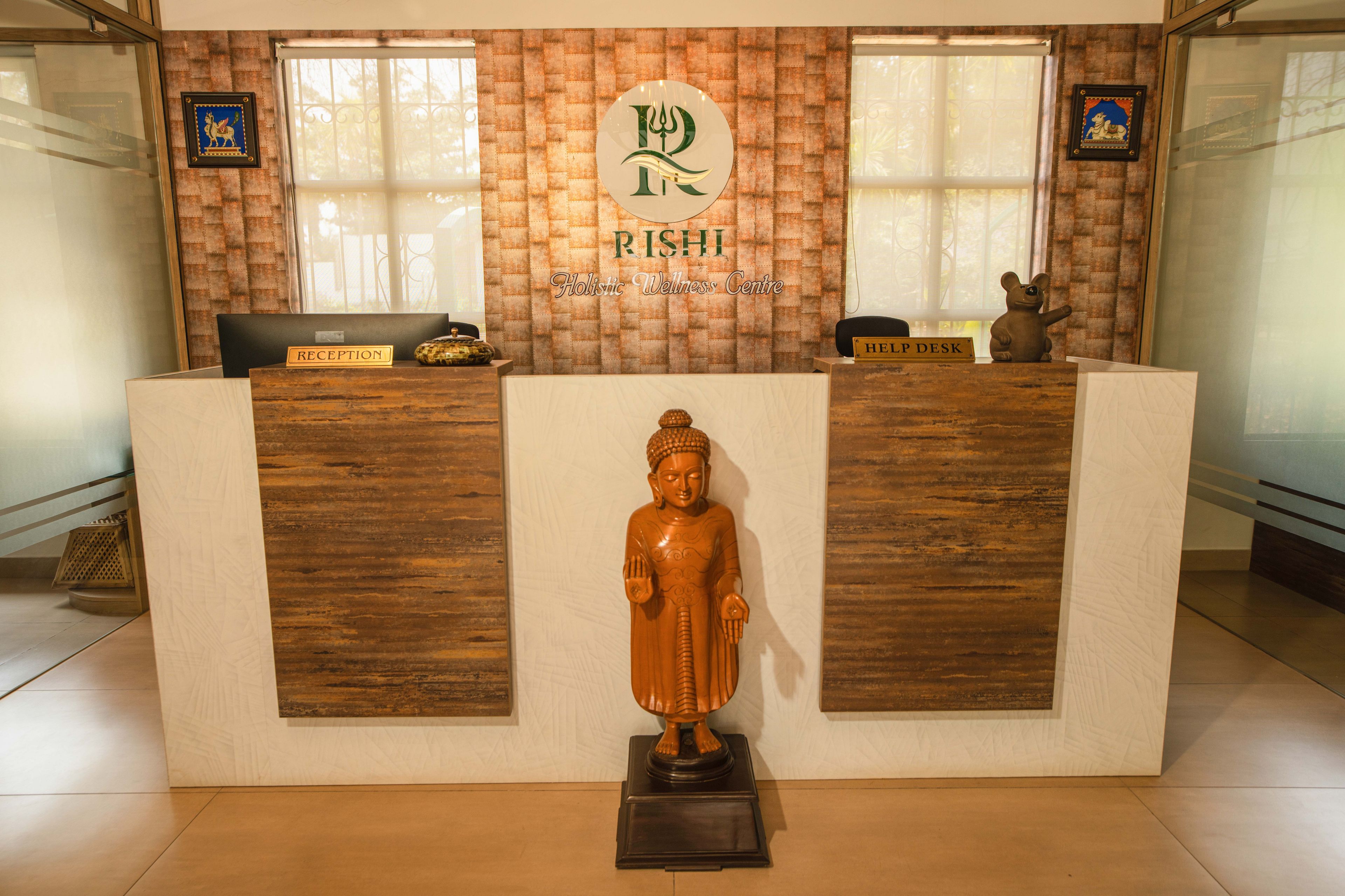Un mostrador de recepción con detalles de madera, una estatua de Buda al frente y dos ventanas con cortinas.