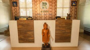 Un mostrador de recepción con detalles de madera, una estatua de Buda al frente y dos ventanas con cortinas.