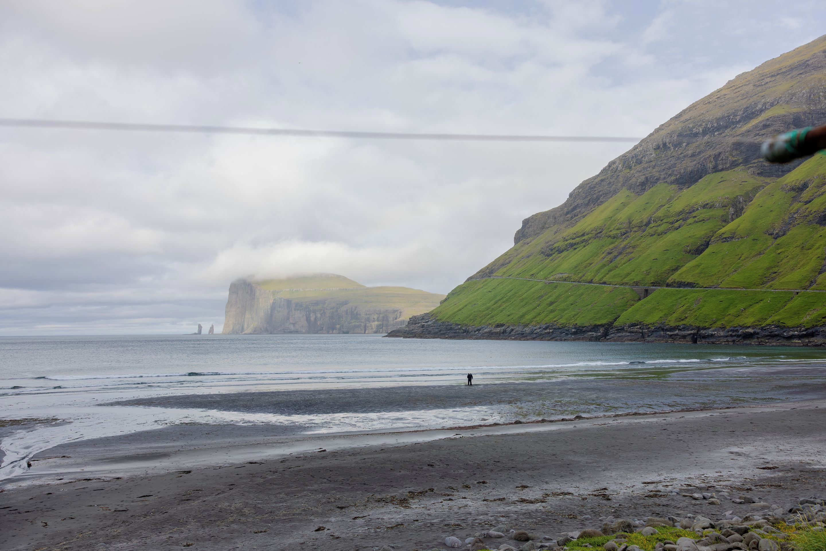 Un paisaje costero con una playa rocosa, una persona de pie en la orilla y una montaña al fondo.