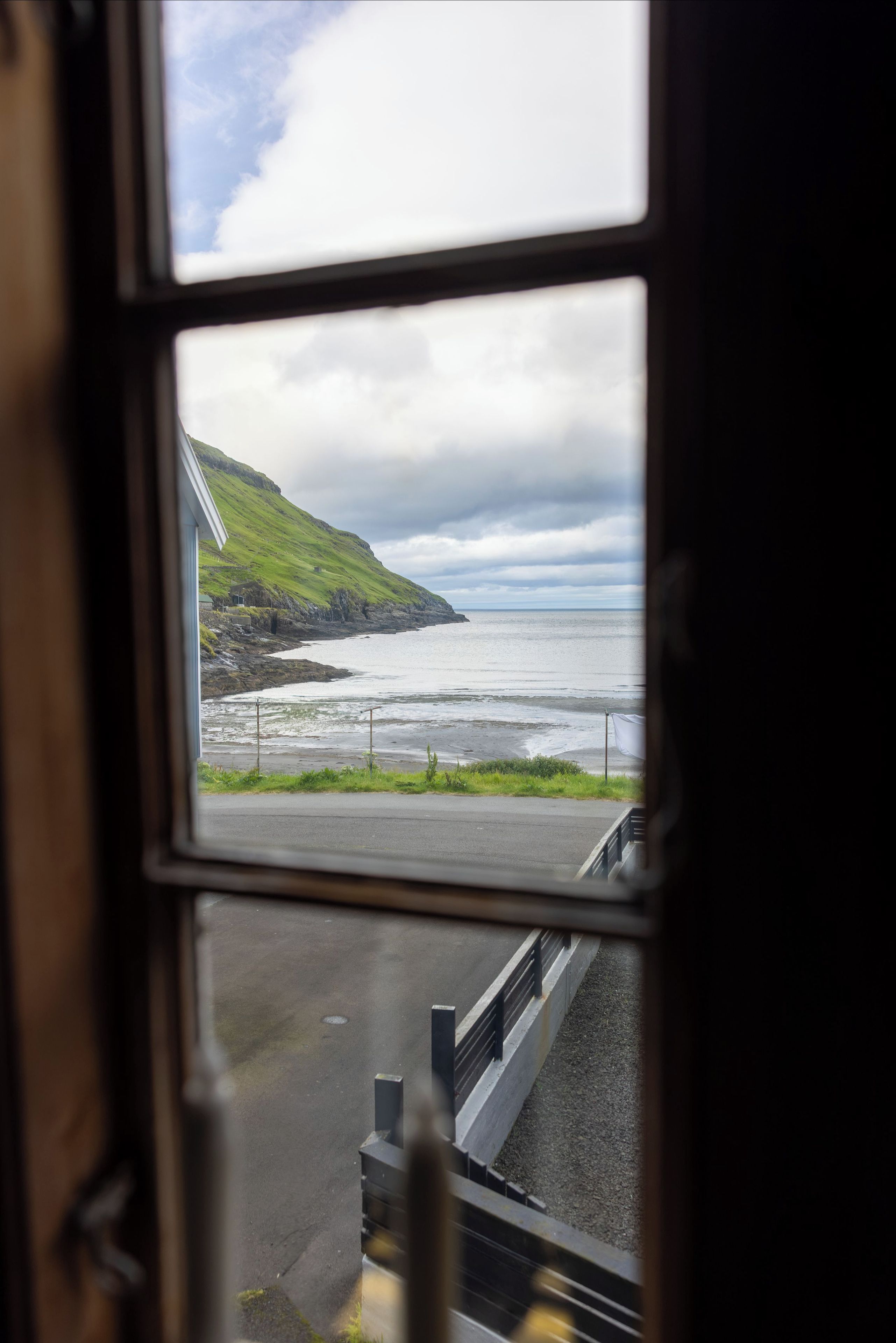 Blick aufs Meer durch ein Fenster, mit einer grünen Hügelkuppe und einer Straße, die zum Meer führt.