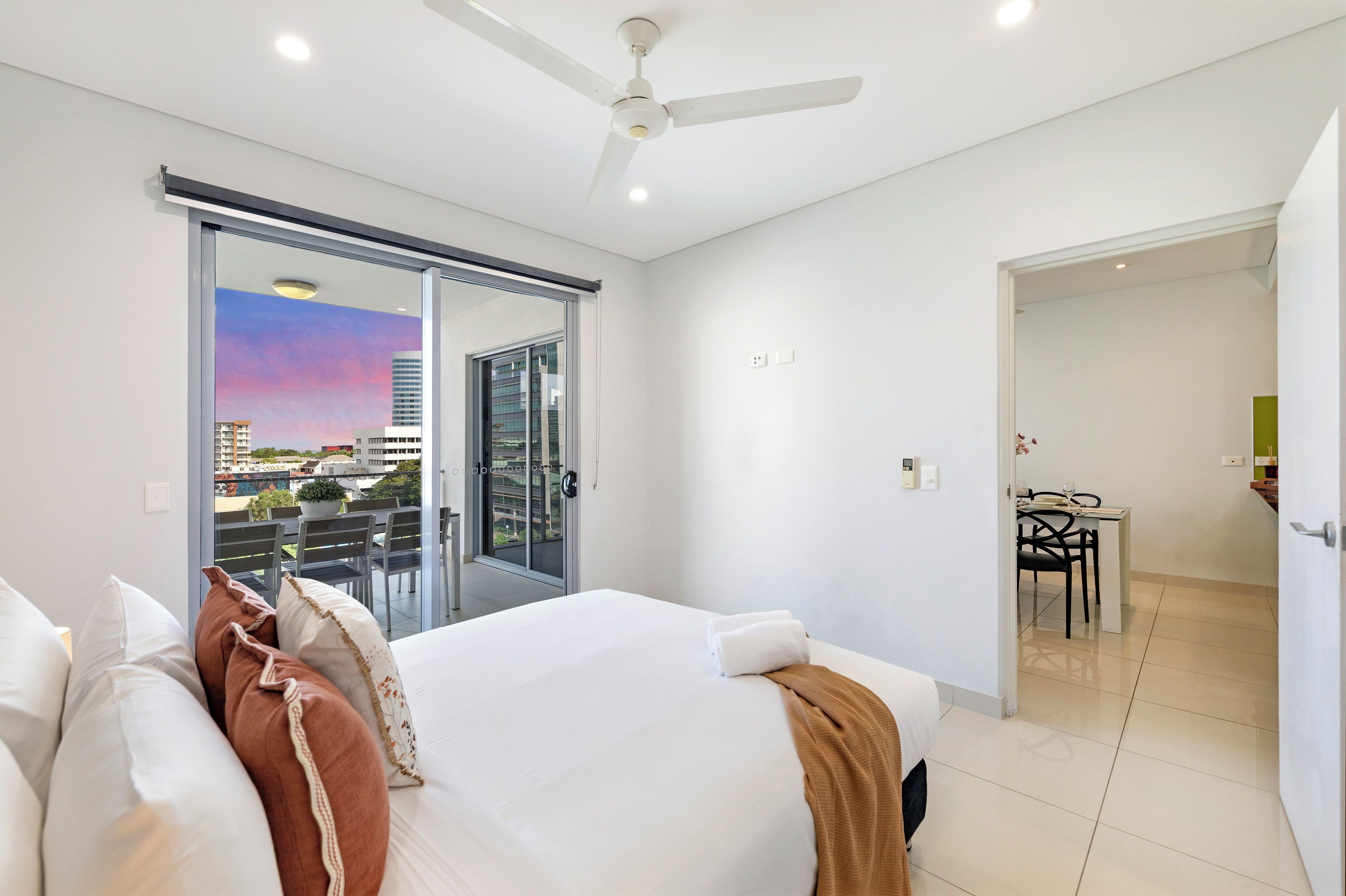 A bedroom with a bed, a ceiling fan, a sliding glass door to a balcony, and a view of the cityscape.