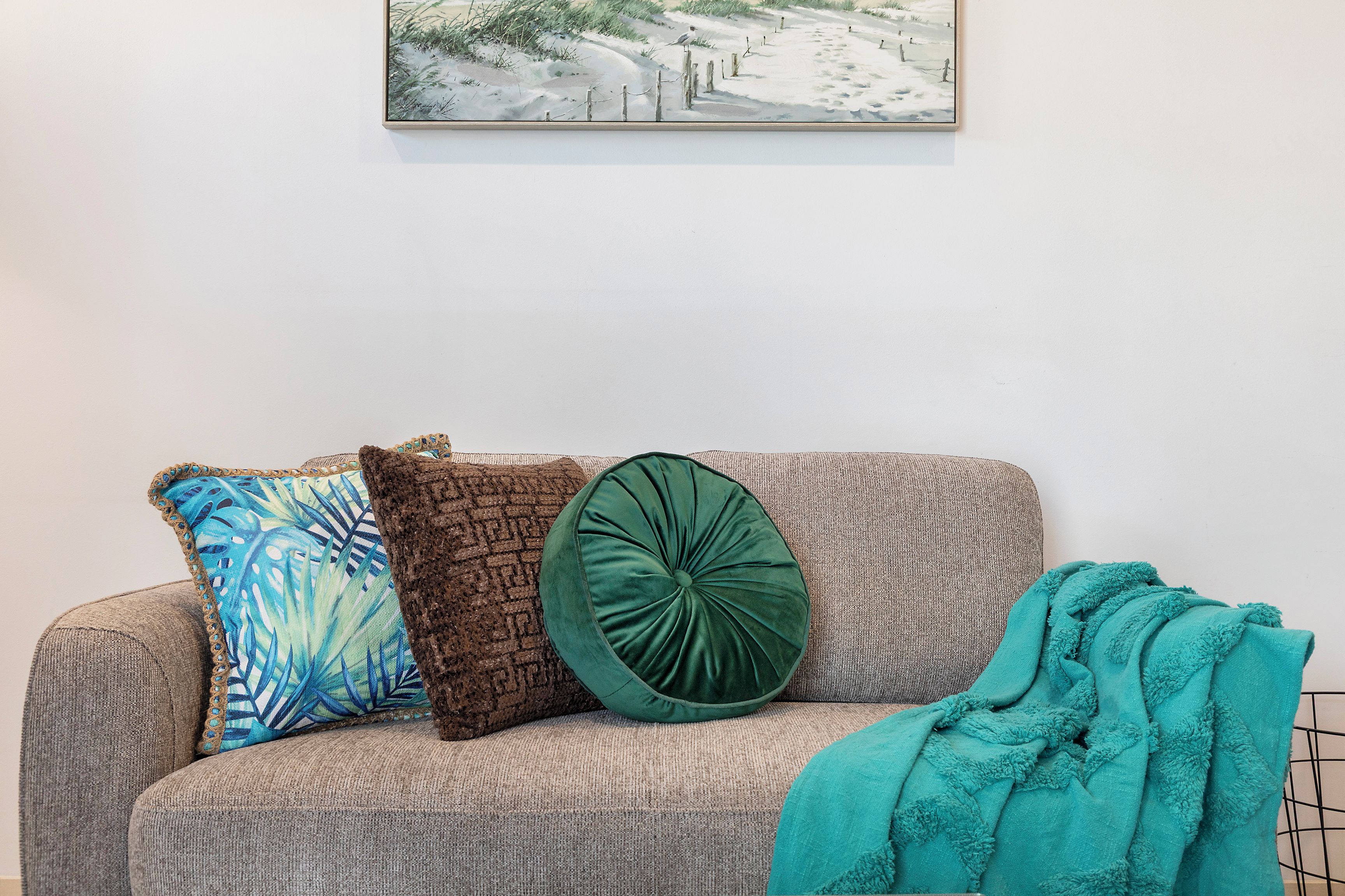A sofa with patterned cushions and a green decorative pillow, a teal throw blanket, and a painting on the wall.