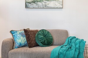 Un sofà amb coixins estampats i un coixí decoratiu verd, una manta color turquesa, i un quadre a la paret.