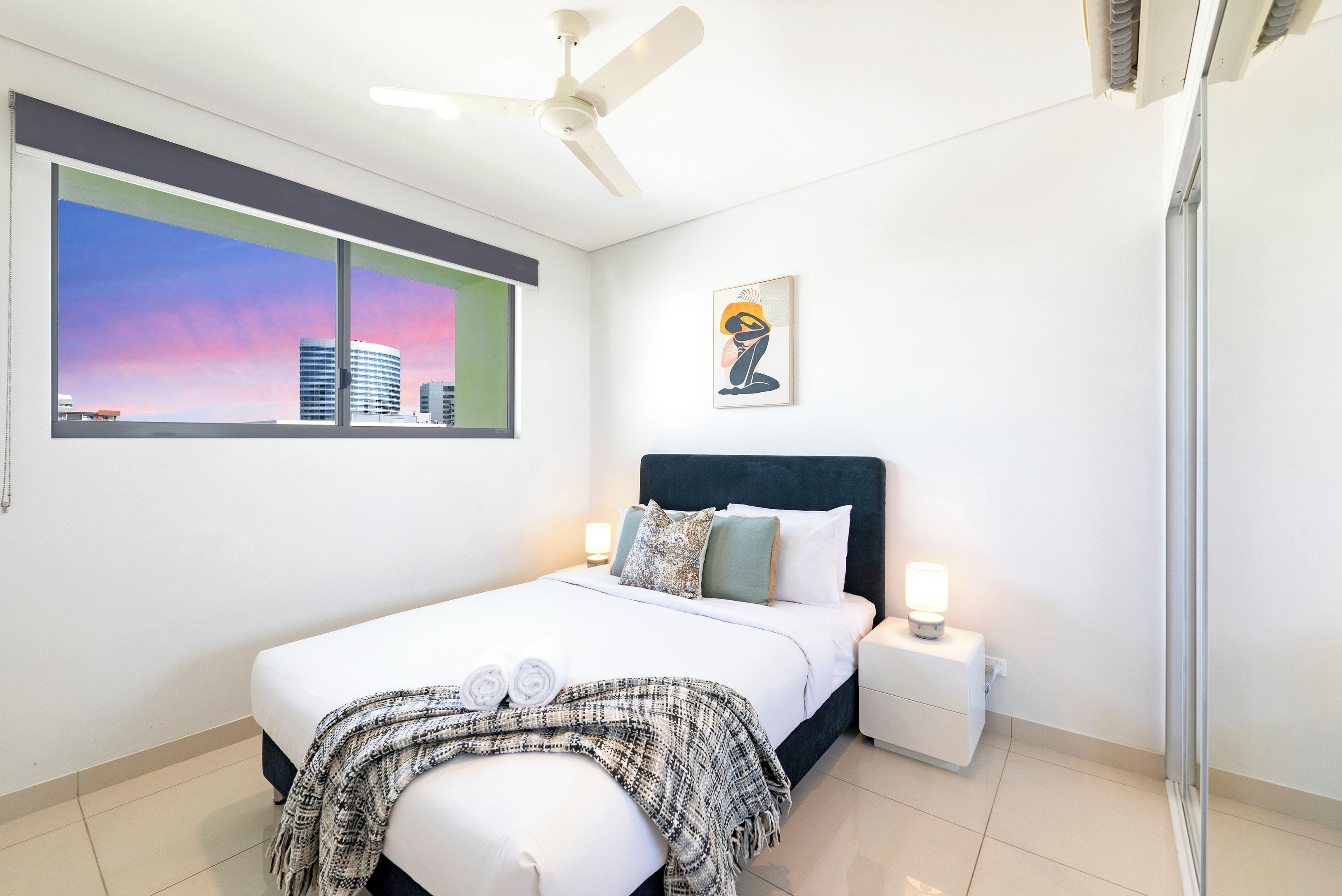 Une chambre d’hôtel avec un lit, une table de chevet, une lampe, un ventilateur de plafond, une fenêtre donnant sur le paysage urbain et un tableau au mur.