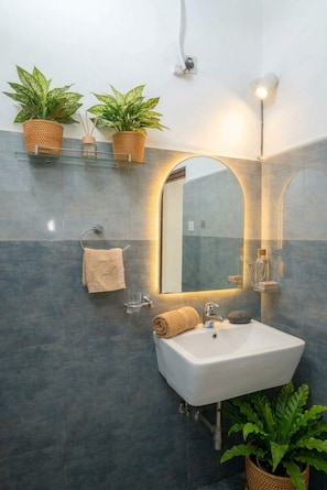 A bathroom with a white sink, two potted plants on a shelf, a towel rack, and a mirror with a light above it.